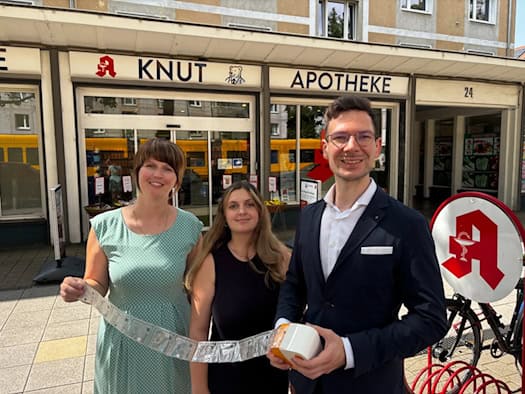 Romy Schneider, Jasmin Schmähl und Michael Gäbe stehen vor dem Eingang der Knut-Apotheke in Dresden und präsentieren ein langes Band maschinell verblisterter Medikamentenbeutel.