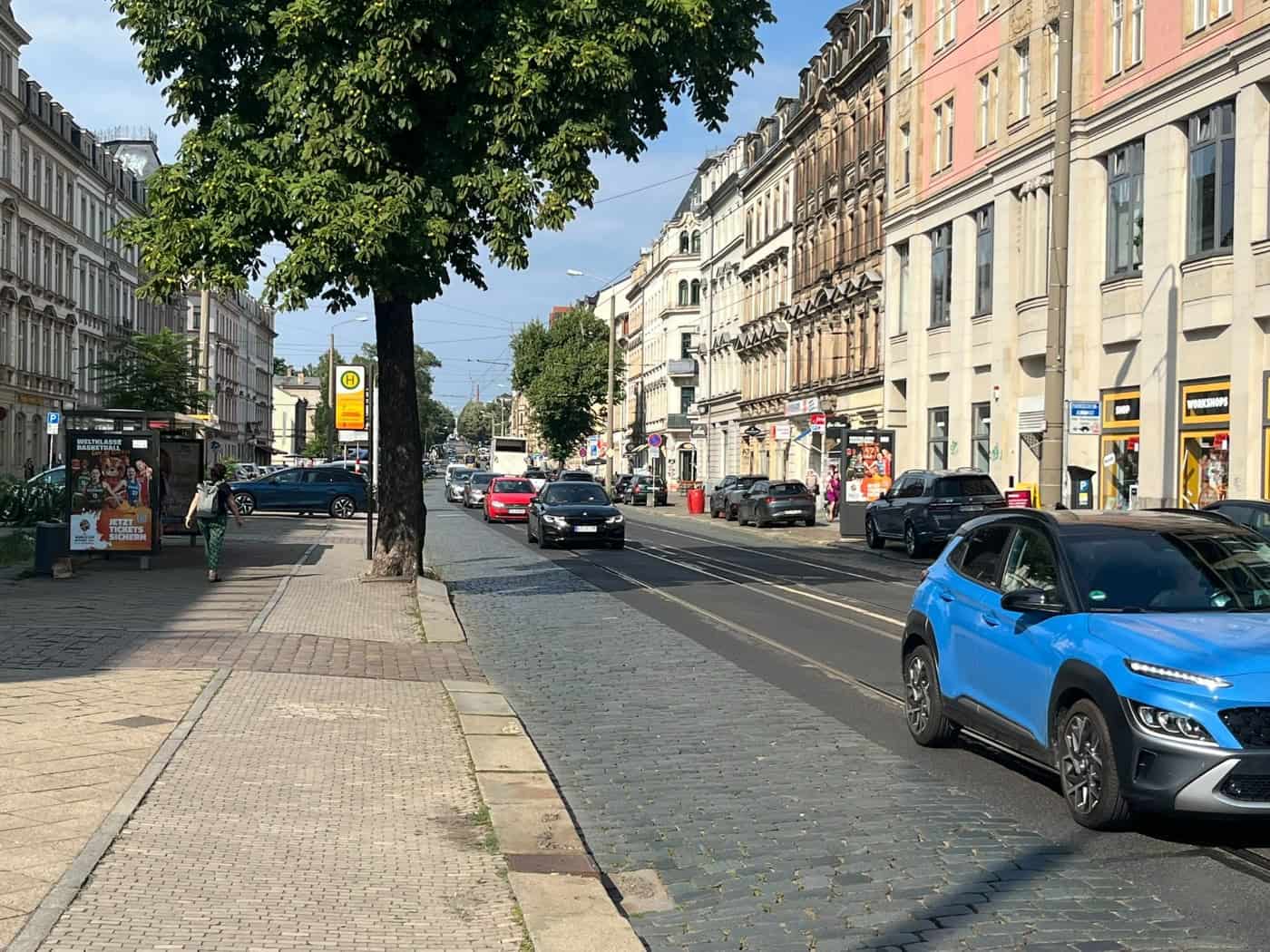 Straßenzug mit Kopfsteinpflaster und Straßenbahnschienen, gesäumt von Altbauten. Autos fahren auf der Fahrbahn, links befindet sich eine Tramhaltestelle unter einem grünen Baum.