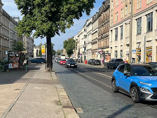 Straßenzug mit Kopfsteinpflaster und Straßenbahnschienen, gesäumt von Altbauten. Autos fahren auf der Fahrbahn, links befindet sich eine Tramhaltestelle unter einem grünen Baum.