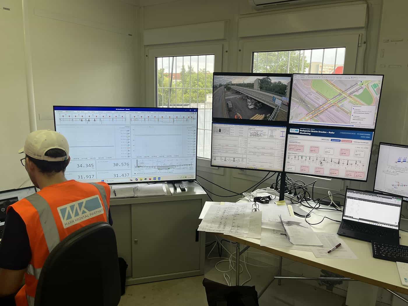 Blick von hinten auf einen Mitarbeiter mit orangefarbener Warnweste und Mütze in einer Kommandozentrale. Vor ihm befinden sich mehrere große Bildschirme mit Live-Kamerabildern der Brücke, Karten und Messwert-Dashboards zur Überwachung.