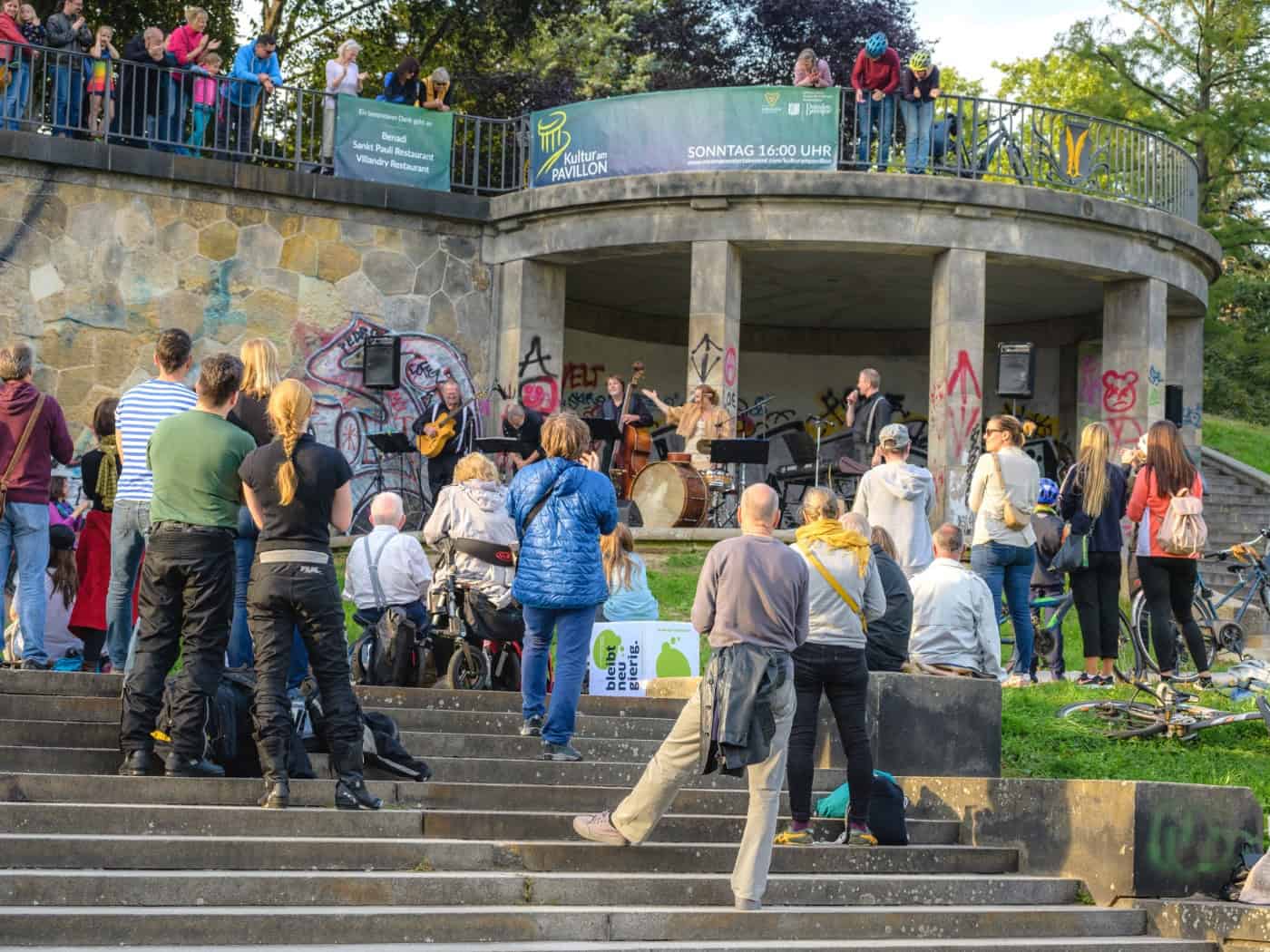 Publikum steht und sitzt auf der Freitreppe vor dem runden Steinpavillon an der Dresdner Albertbrücke. Eine Band mit Kontrabass und Schlagzeug tritt im überdachten Säulenbau auf, Zuschauer lehnen auch oben an der Balustrade.