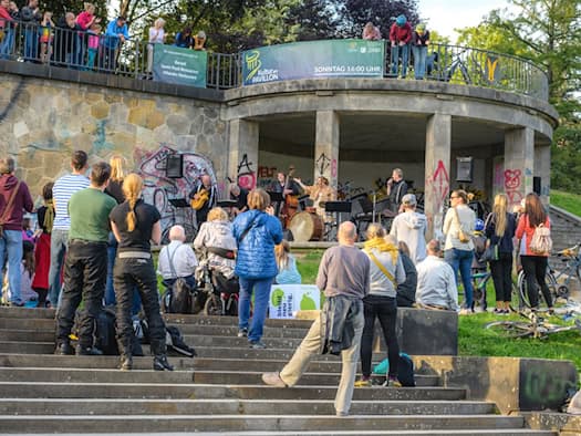 Publikum steht und sitzt auf der Freitreppe vor dem runden Steinpavillon an der Dresdner Albertbrücke. Eine Band mit Kontrabass und Schlagzeug tritt im überdachten Säulenbau auf, Zuschauer lehnen auch oben an der Balustrade.