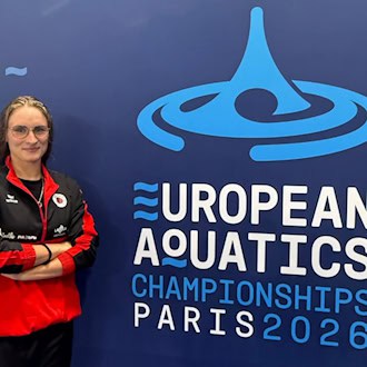 Schwimmerin Maya Tobehn steht lächelnd mit verschränkten Armen und Brille vor einer blauen Wand mit dem Logo der European Aquatics Championships Paris 2026. Sie trägt eine rot-schwarze Trainingsjacke des Dresdner SC.