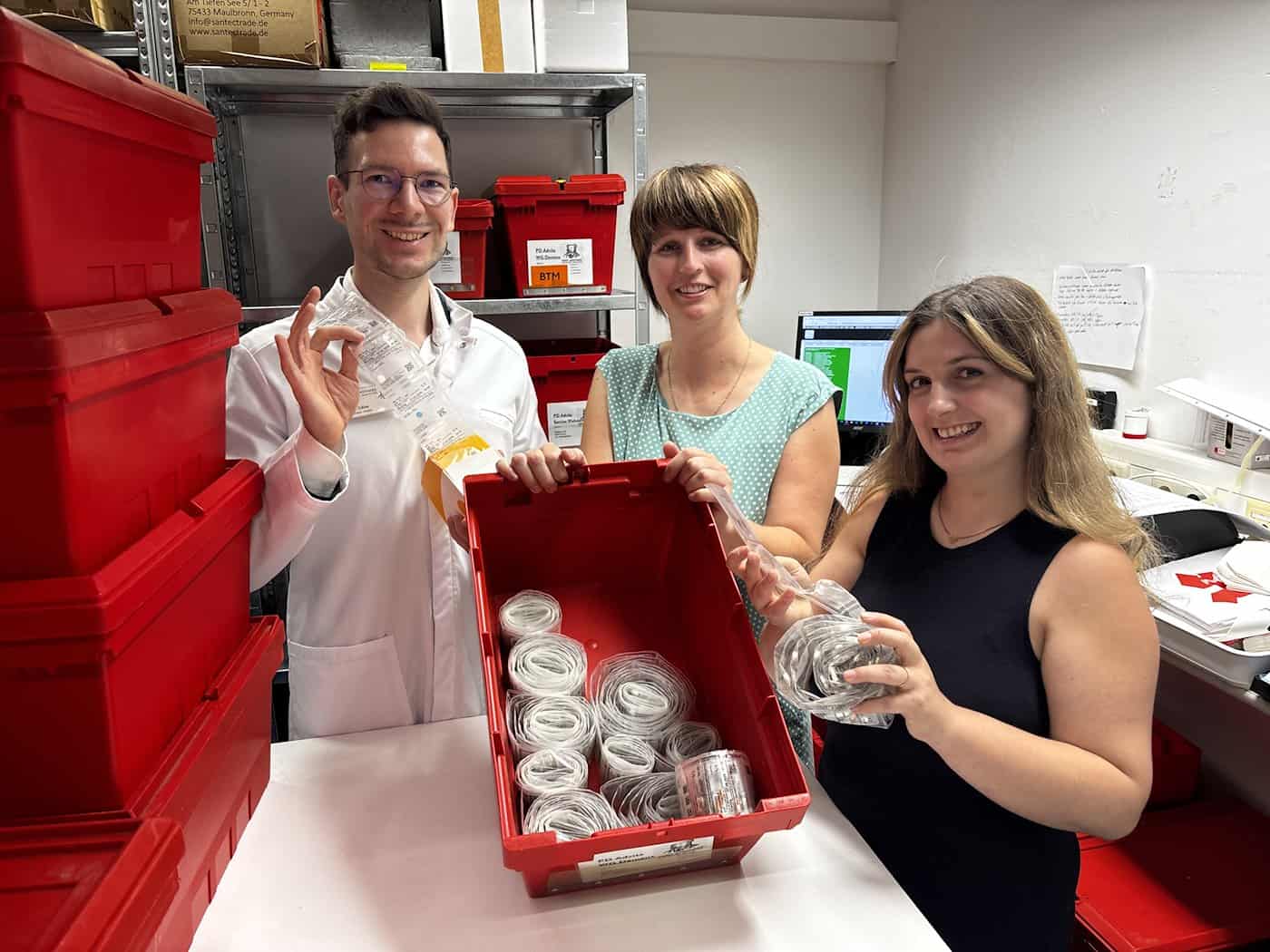 Michael Gäbe, Romy Schneider und Jasmin Schmähl stehen im Vorbereitungsraum der Apotheke vor roten Sortierkisten und halten verblisterte Medikamentenbeutel in den Händen.  Bildunterschrift (246 Zeichen)