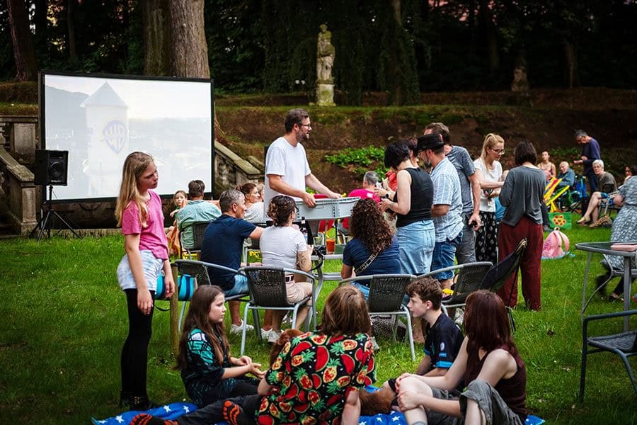 Zahlreiche Menschen aller Alter sitzen auf Decken und Stühlen auf einer Wiese und interagieren miteinander, im Hintergrund eine Leinwand zur Filmprojektion.