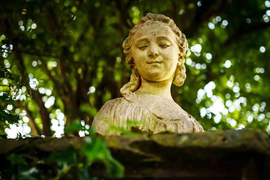 Kopf- und Schulterbereich einer Plastik aus Stein, die eine Dame oder Mädchen darstellt, umgegeben von (von der Kamera nicht fokussierter und damit unscharfer) grüner Natur.
