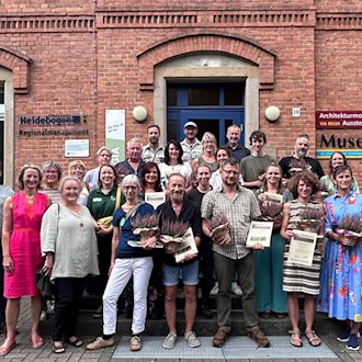 Gruppenfoto von rund 20 Menschen vor den Stufen eines roten Klinkergebäudes. Mehrere Frauen und Männer halten gedruckte Zertifikate und kleine, blühende Heidekraut-Pflanzen in den Händen und lächeln in die Kamera.