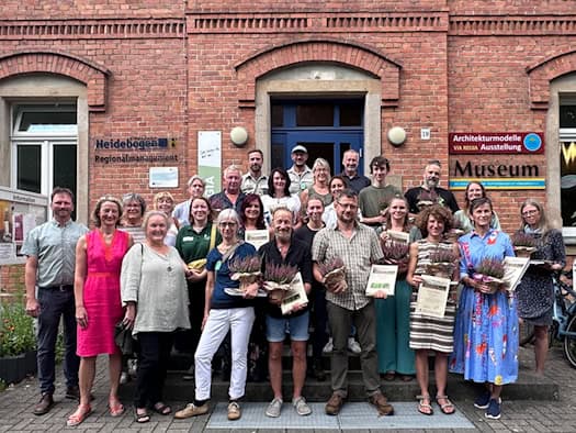 Gruppenfoto von rund 20 Menschen vor den Stufen eines roten Klinkergebäudes. Mehrere Frauen und Männer halten gedruckte Zertifikate und kleine, blühende Heidekraut-Pflanzen in den Händen und lächeln in die Kamera.