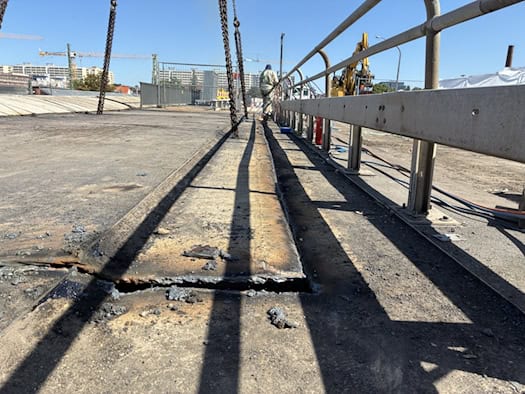 Nahaufnahme der Fahrbahn der Nossener Brücke in Dresden bei Sonnenschein. Ein ausgeschnittenes Beton- und Stahlsegment des Kragarms hängt an Kranketten. Im Hintergrund steht ein Bauarbeiter am Brückengeländer.