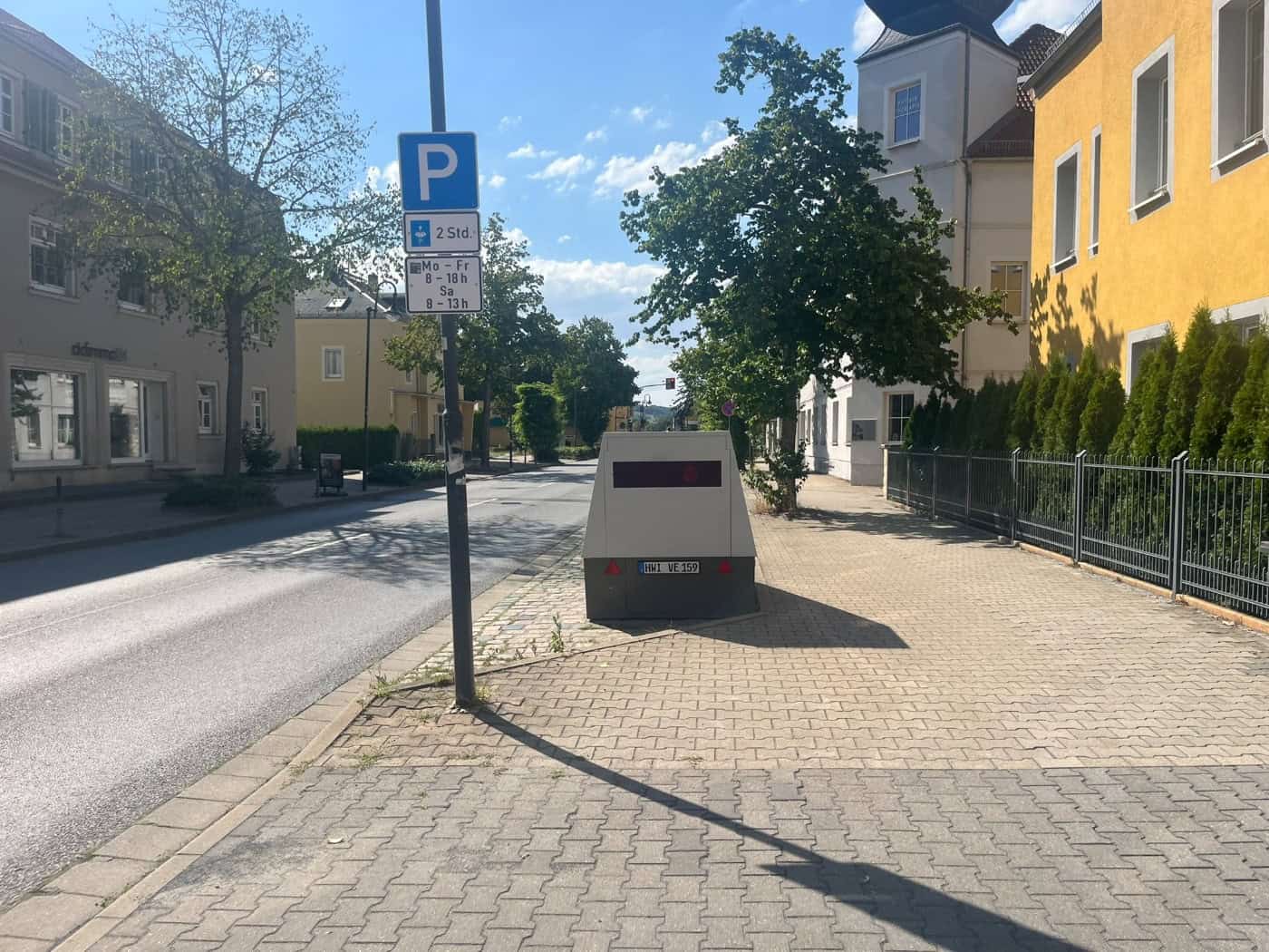 Blick entlang einer sonnenbeschienenen Hauptstraße mit Gehweg und Parkplatzschild in Dresden-Cossebaude. Auf den Seiten stehen Wohnhäuser und Bäume unter leicht bewölktem blauem Himmel.