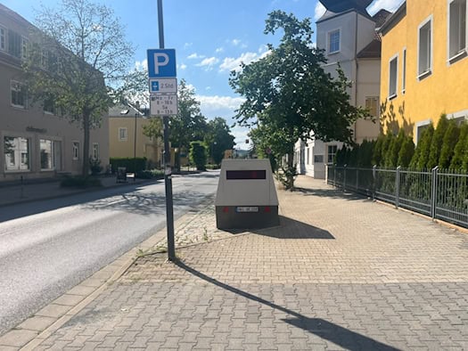 Blick entlang einer sonnenbeschienenen Hauptstraße mit Gehweg und Parkplatzschild in Dresden-Cossebaude. Auf den Seiten stehen Wohnhäuser und Bäume unter leicht bewölktem blauem Himmel.