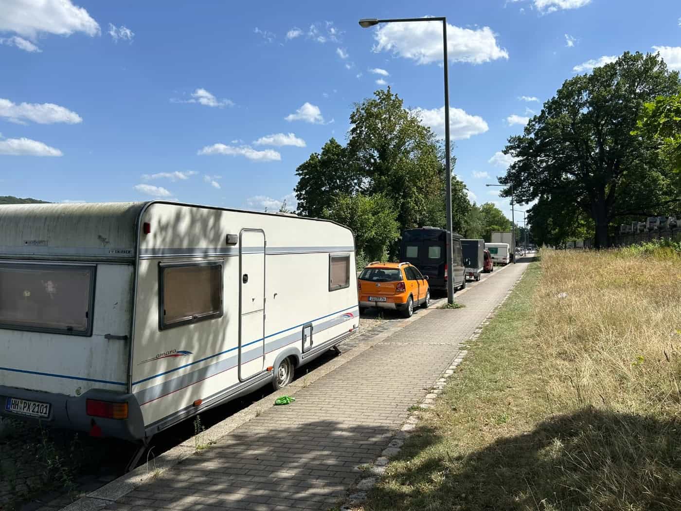 Ein großer weißer Wohnwagen parkt auf dem Randstreifen an der Tolkewitzer Straße in Dresden. Dahinter stehen ein orangener Pkw und weitere Transporter aufgereiht an einem Gehweg unter blauem Sommerhimmel.