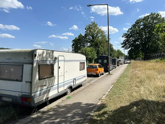 Ein großer weißer Wohnwagen parkt auf dem Randstreifen an der Tolkewitzer Straße in Dresden. Dahinter stehen ein orangener Pkw und weitere Transporter aufgereiht an einem Gehweg unter blauem Sommerhimmel.