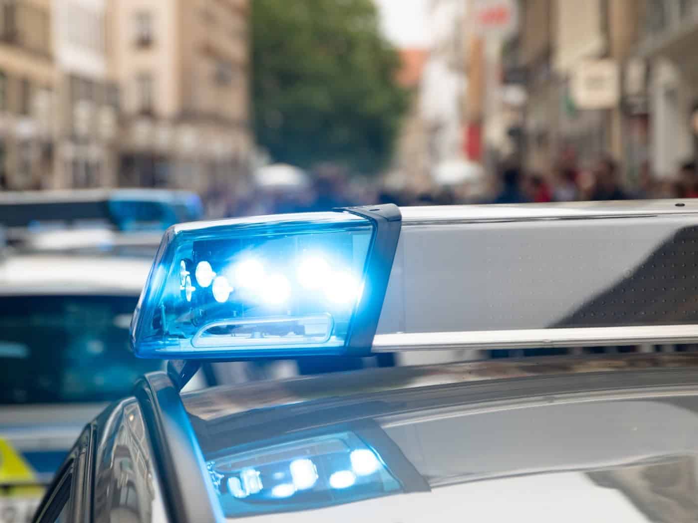 Nahaufnahme eines eingeschalteten Blaulichts auf dem Dach eines Streifenwagens der Polizei. Im unscharfen Hintergrund steht ein weiteres Einsatzfahrzeug in einer belebten Stadtstraße.