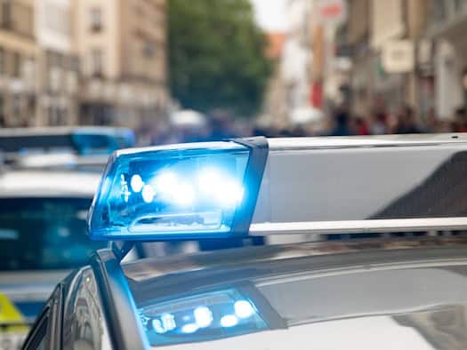 Nahaufnahme eines eingeschalteten Blaulichts auf dem Dach eines Streifenwagens der Polizei. Im unscharfen Hintergrund steht ein weiteres Einsatzfahrzeug in einer belebten Stadtstraße.