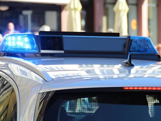 Symbolfoto: Nahaufnahme vom Dach eines Polizeifahrzeugs mit eingeschaltetem, leuchtendem Blaulicht auf der Lichtleiste vor unscharfem Stadtkulissen-Hintergrund.