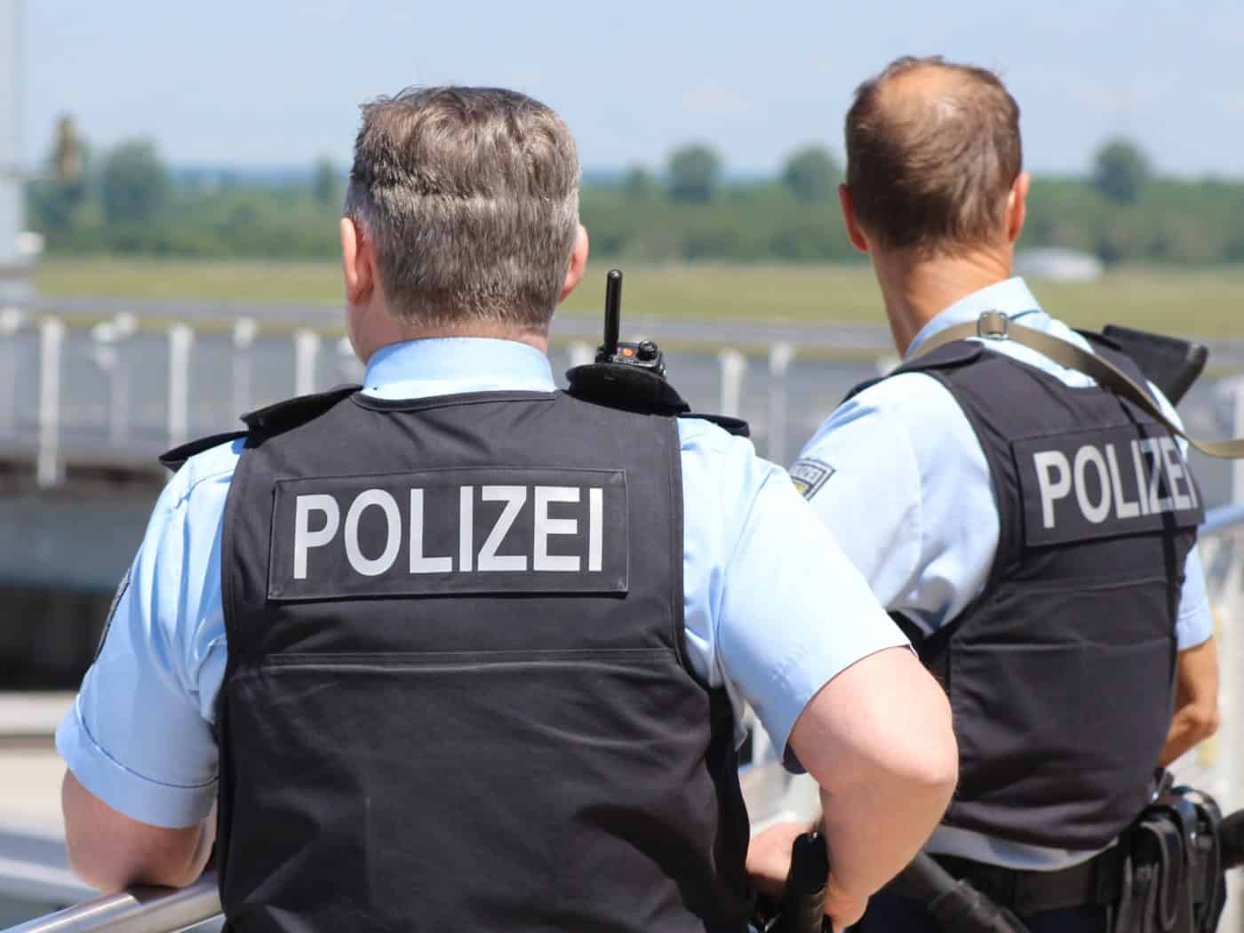Rückansicht von zwei Polizeibeamten in Kurzarmhemden und dunklen Schutzwesten mit der Aufschrift „POLIZEI“. Sie stehen an einem Geländer im Außenbereich.