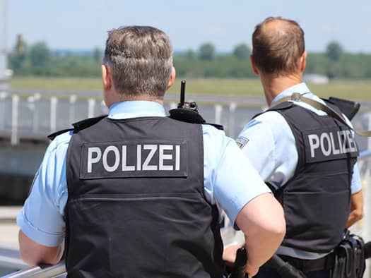 Rückansicht von zwei Polizeibeamten in Kurzarmhemden und dunklen Schutzwesten mit der Aufschrift „POLIZEI“. Sie stehen an einem Geländer im Außenbereich.