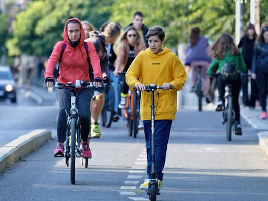 Ein Junge in gelbem Hoodie auf einem E-Scooter und eine Frau in roter Jacke auf dem Fahrrad fahren nebeneinander auf einer abgegrenzten Radspur in der Stadt. Im Hintergrund sind weitere Radfahrende und Passanten zu sehen.