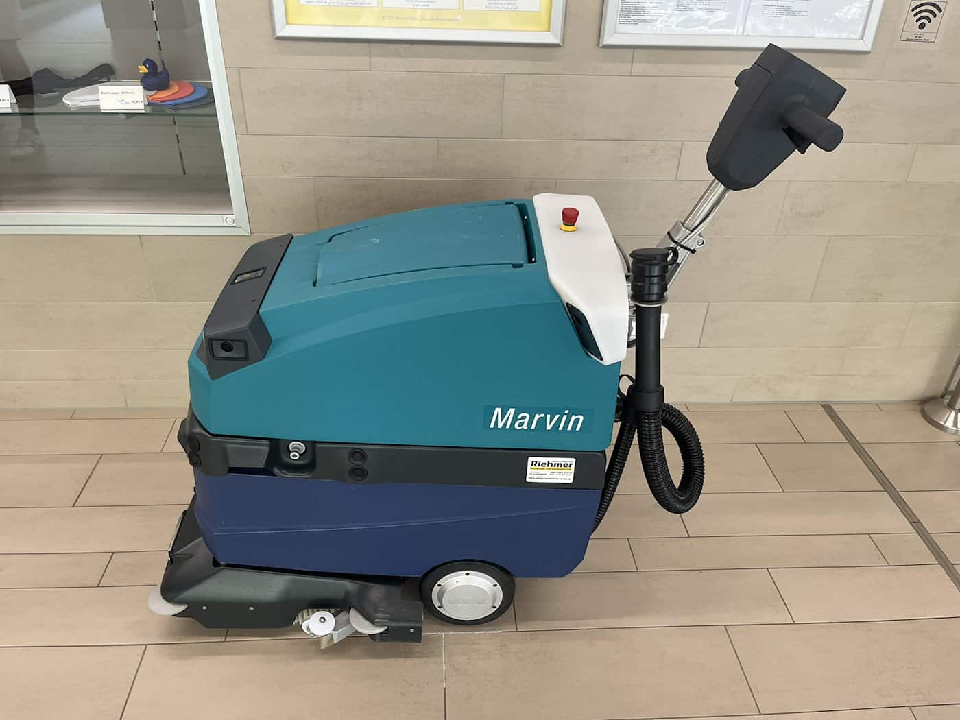 Ein türkis-blauer Scheuersaug-Reinigungsroboter mit der Aufschrift 'Marvin' steht auf hell gefliestem Boden vor einer Glaswand.