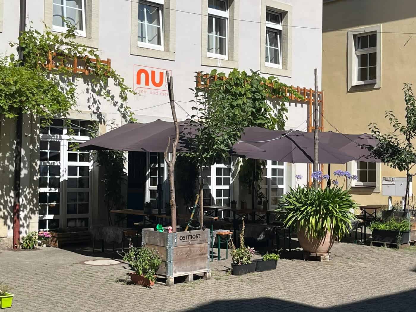Pflasterfreisitz im begrünten Hinterhof des Restaurants nu! in Dresden-Neustadt. Unter großen dunklen Sonnenschirmen stehen Holzgarnituren, umgeben von Pflanzkübeln mit Blumen und kletterndem Wein an der hellen Hauswand.