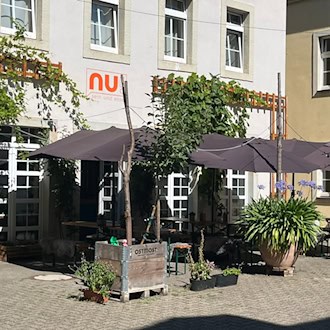 Pflasterfreisitz im begrünten Hinterhof des Restaurants nu! in Dresden-Neustadt. Unter großen dunklen Sonnenschirmen stehen Holzgarnituren, umgeben von Pflanzkübeln mit Blumen und kletterndem Wein an der hellen Hauswand.