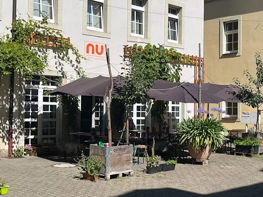 Pflasterfreisitz im begrünten Hinterhof des Restaurants nu! in Dresden-Neustadt. Unter großen dunklen Sonnenschirmen stehen Holzgarnituren, umgeben von Pflanzkübeln mit Blumen und kletterndem Wein an der hellen Hauswand.
