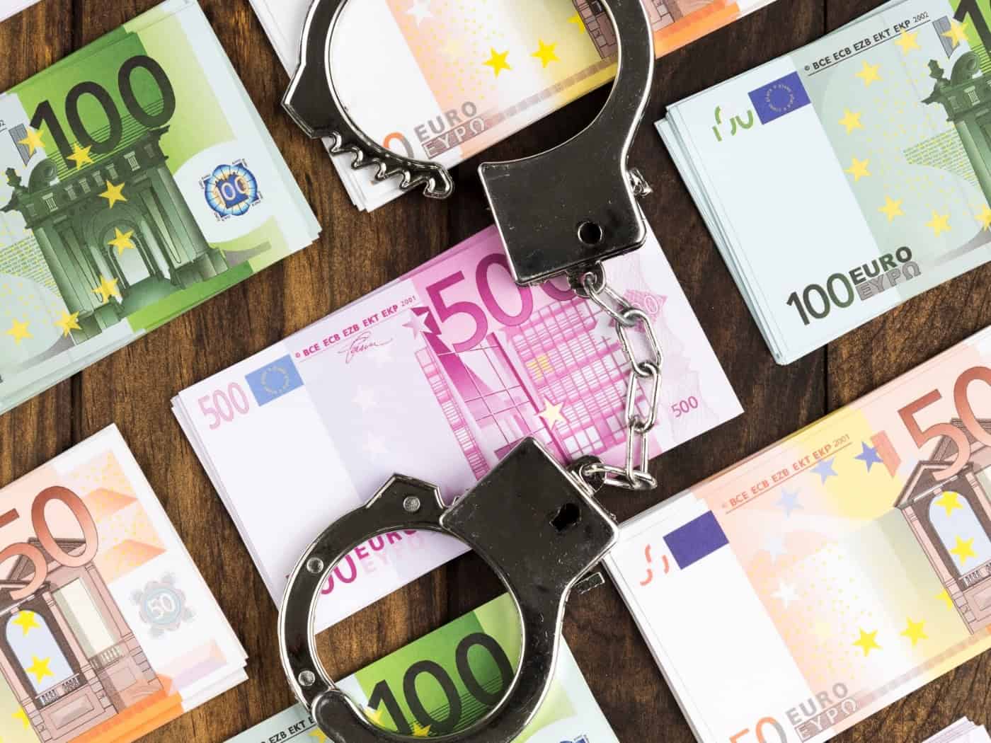 Flatlay-Aufnahme auf einem dunklen Holztisch: Mehrere ordentlich gestapelte Bündel von 50-, 100- und 500-Euro-Geldscheinen liegen verteilt, in der Mitte liegt eine geöffnete silberne Handschelle.