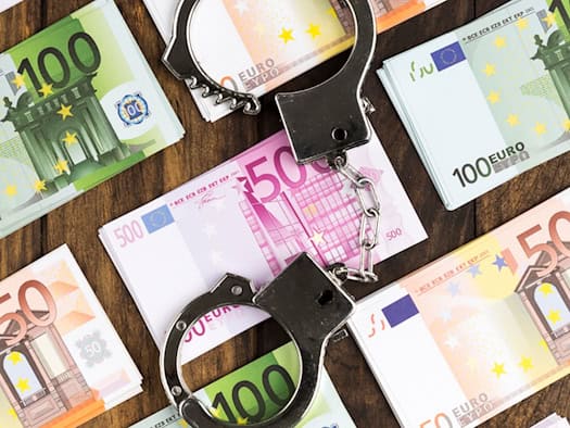 Flatlay-Aufnahme auf einem dunklen Holztisch: Mehrere ordentlich gestapelte Bündel von 50-, 100- und 500-Euro-Geldscheinen liegen verteilt, in der Mitte liegt eine geöffnete silberne Handschelle.