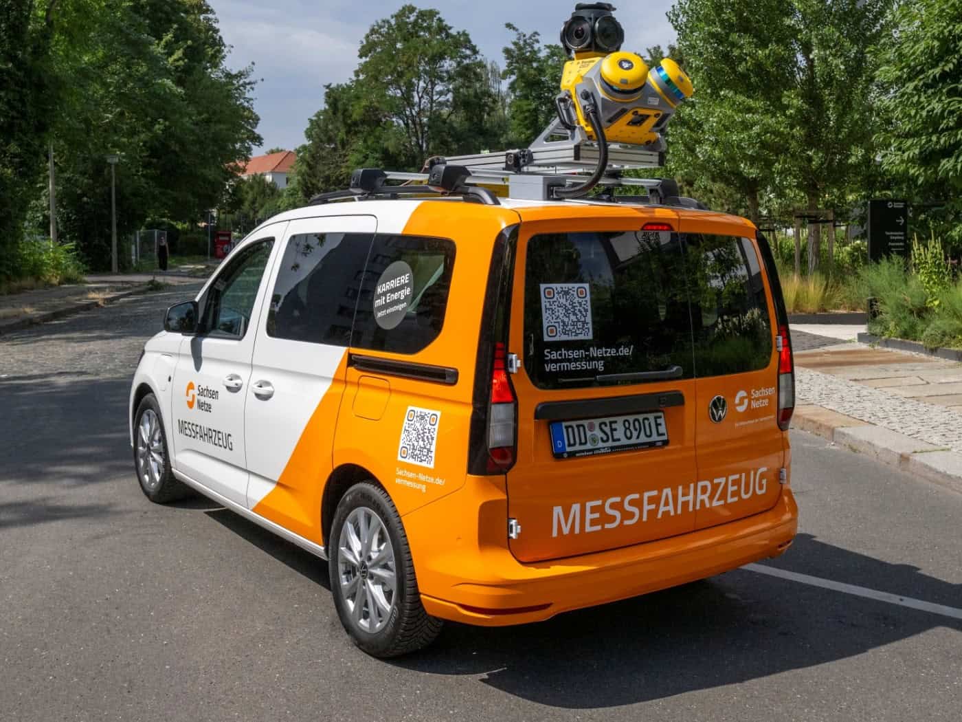 Schrägansicht von hinten auf ein weiß-orangenes Messfahrzeug der SachsenNetze mit gelben 3D-Kameras und Laserscannern auf dem Dach. Das Auto fährt auf einer asphaltierten Straße an einem modernen Gebäude und Bäumen vorbei.