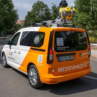 Schrägansicht von hinten auf ein weiß-orangenes Messfahrzeug der SachsenNetze mit gelben 3D-Kameras und Laserscannern auf dem Dach. Das Auto fährt auf einer asphaltierten Straße an einem modernen Gebäude und Bäumen vorbei.