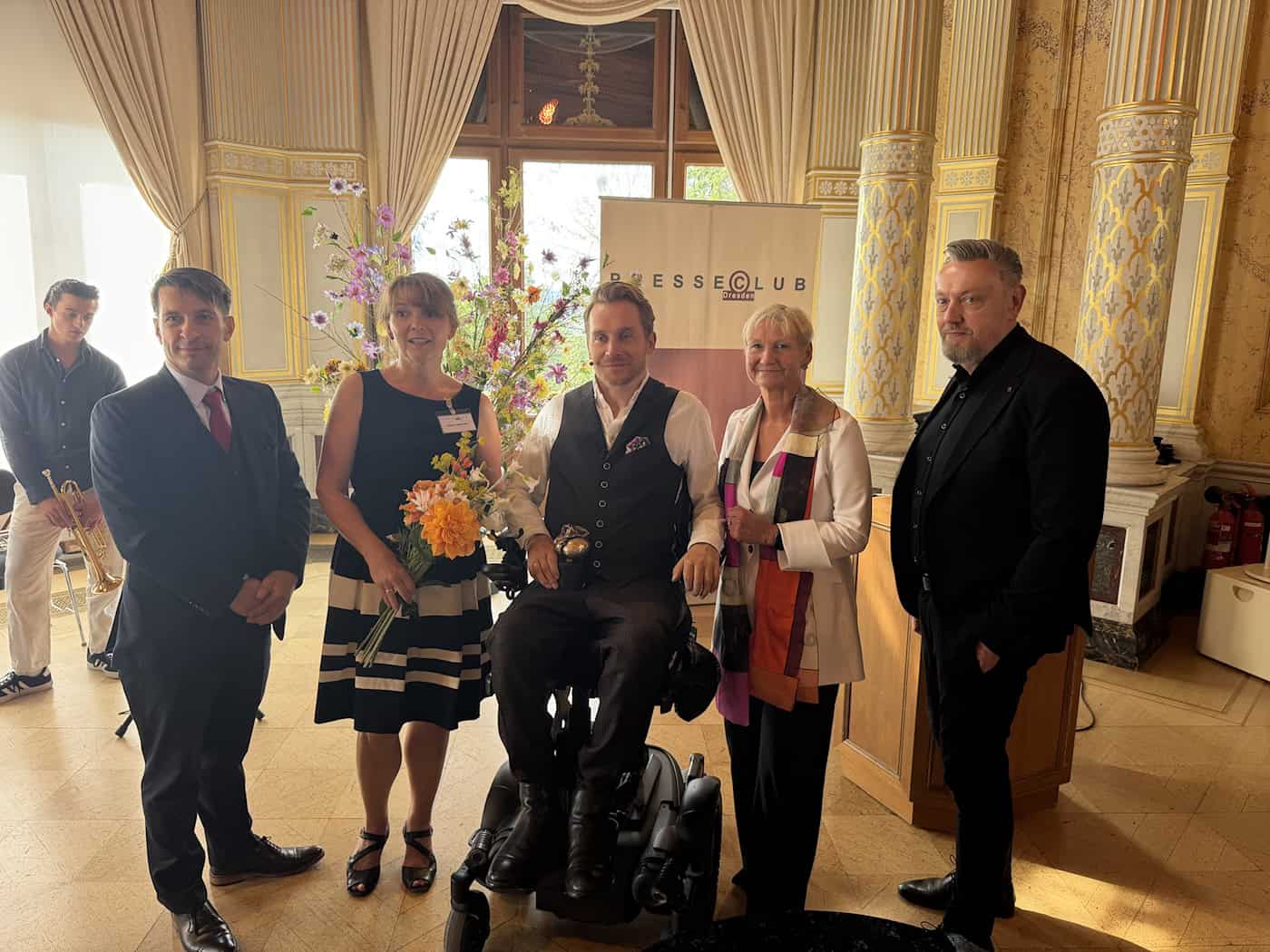 Samuel Koch sitzt mit der Bronze-Trophäe im Rollstuhl im Festsaal von Schloss Albrechtsberg. Er wird eingerahmt von Tobias Wolf, Sabine Mutschke mit Blumenstrauß, Bischöfin Kirsten Fehrs und Andreas Weller, die neben ihm stehen.