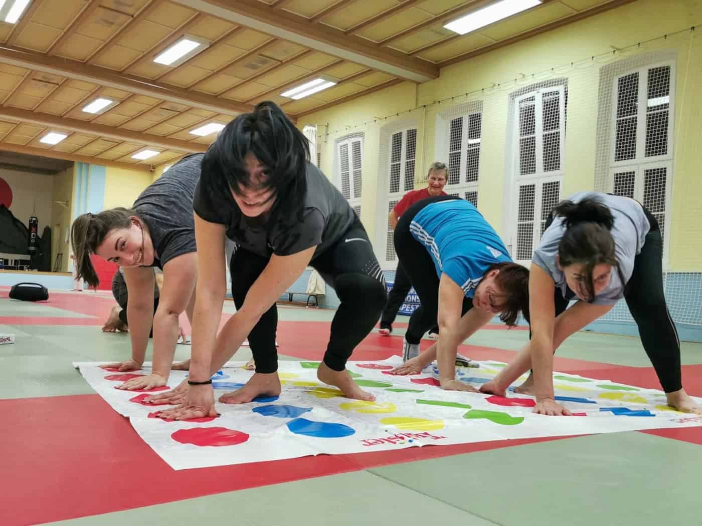 Vier Frauen verrenken sich am Boden auf einer Matte mit farbigen Punkten (vom Spiel "Twister"), in einer Turnhalle