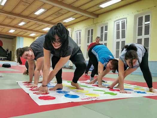Vier Frauen verrenken sich am Boden auf einer Matte mit farbigen Punkten (vom Spiel "Twister"), in einer Turnhalle