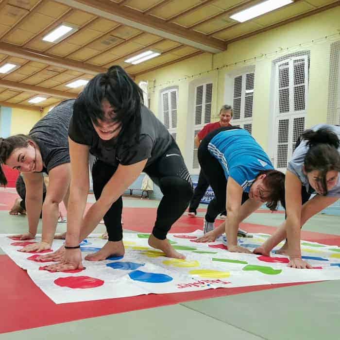 Vier Frauen verrenken sich am Boden auf einer Matte mit farbigen Punkten (vom Spiel "Twister"), in einer Turnhalle
