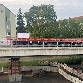 Seitliche Ansicht eines roten, vielachsigen Schwerlasttransporters auf der Brücke Budapester Straße in Dresden. Unter dem Brückenbauwerk sind abgesperrte Notstützen aus Stahl und Betonblöcken zu sehen.