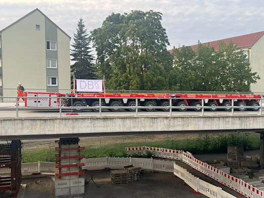 Seitliche Ansicht eines roten, vielachsigen Schwerlasttransporters auf der Brücke Budapester Straße in Dresden. Unter dem Brückenbauwerk sind abgesperrte Notstützen aus Stahl und Betonblöcken zu sehen.
