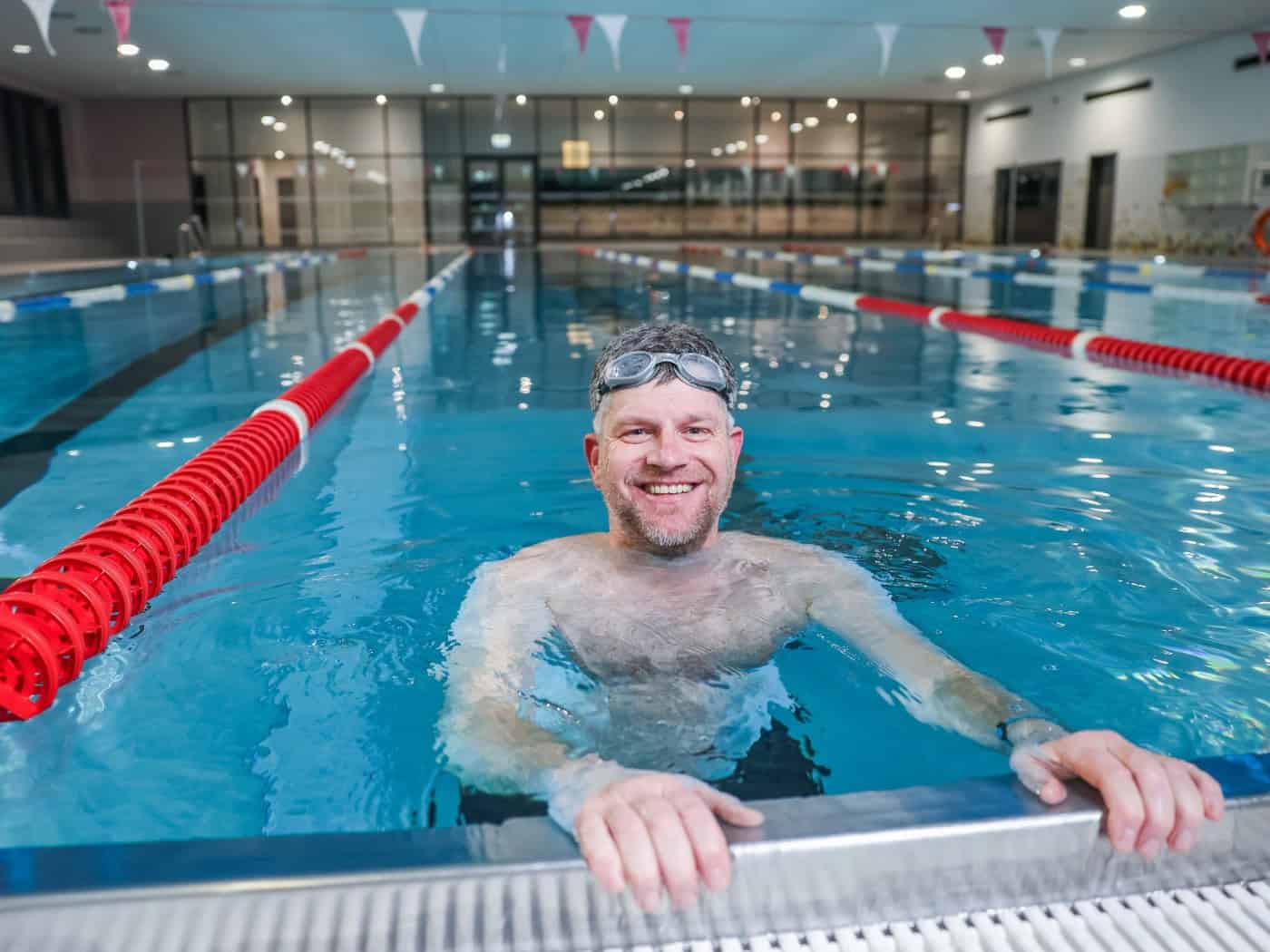 Ein lächelnder Mann mit Schwimmbrille blickt im blau leuchtenden Wasser der Schwimmhalle Bühlau über den Beckenrand. Im Hintergrund sind abgegrenzte Schwimmbahnen mit rot-weißen Leinen zu sehen.