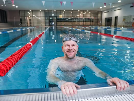 Ein lächelnder Mann mit Schwimmbrille blickt im blau leuchtenden Wasser der Schwimmhalle Bühlau über den Beckenrand. Im Hintergrund sind abgegrenzte Schwimmbahnen mit rot-weißen Leinen zu sehen.