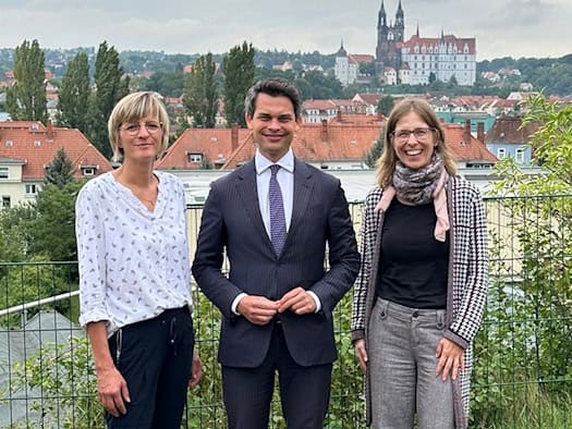 Dr. Christoph Ploß steht im Anzug zwischen Jana Niehoff und Sindy Vogel auf einer Wiese vor einem grünen Zaun. Im Hintergrund ist die Stadt Meißen mit der Albrechtsburg und dem Meißner Dom unter bedecktem Himmel zu sehen.