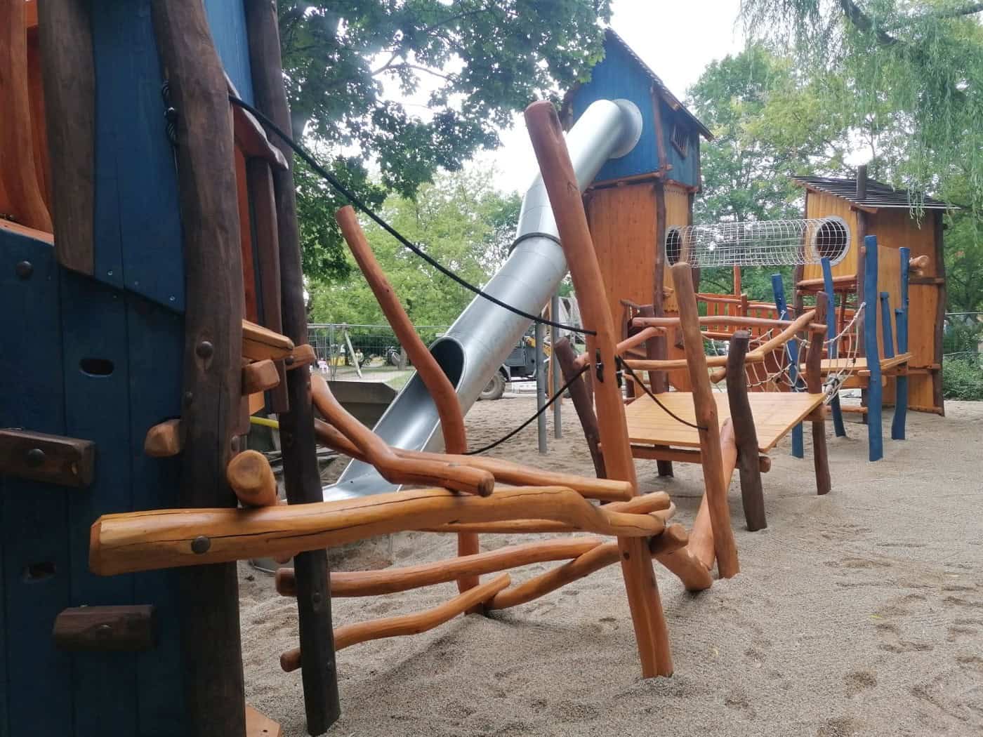 Ein großes Klettergerüst aus Naturholz mit blauen Spielelementen, einer geschwungenen silbernen Röhrenrutsche, Kletterstämmen und einem Netztunnel auf einem Sandspielplatz im Grünen.