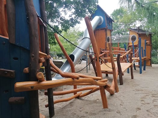 Ein großes Klettergerüst aus Naturholz mit blauen Spielelementen, einer geschwungenen silbernen Röhrenrutsche, Kletterstämmen und einem Netztunnel auf einem Sandspielplatz im Grünen.