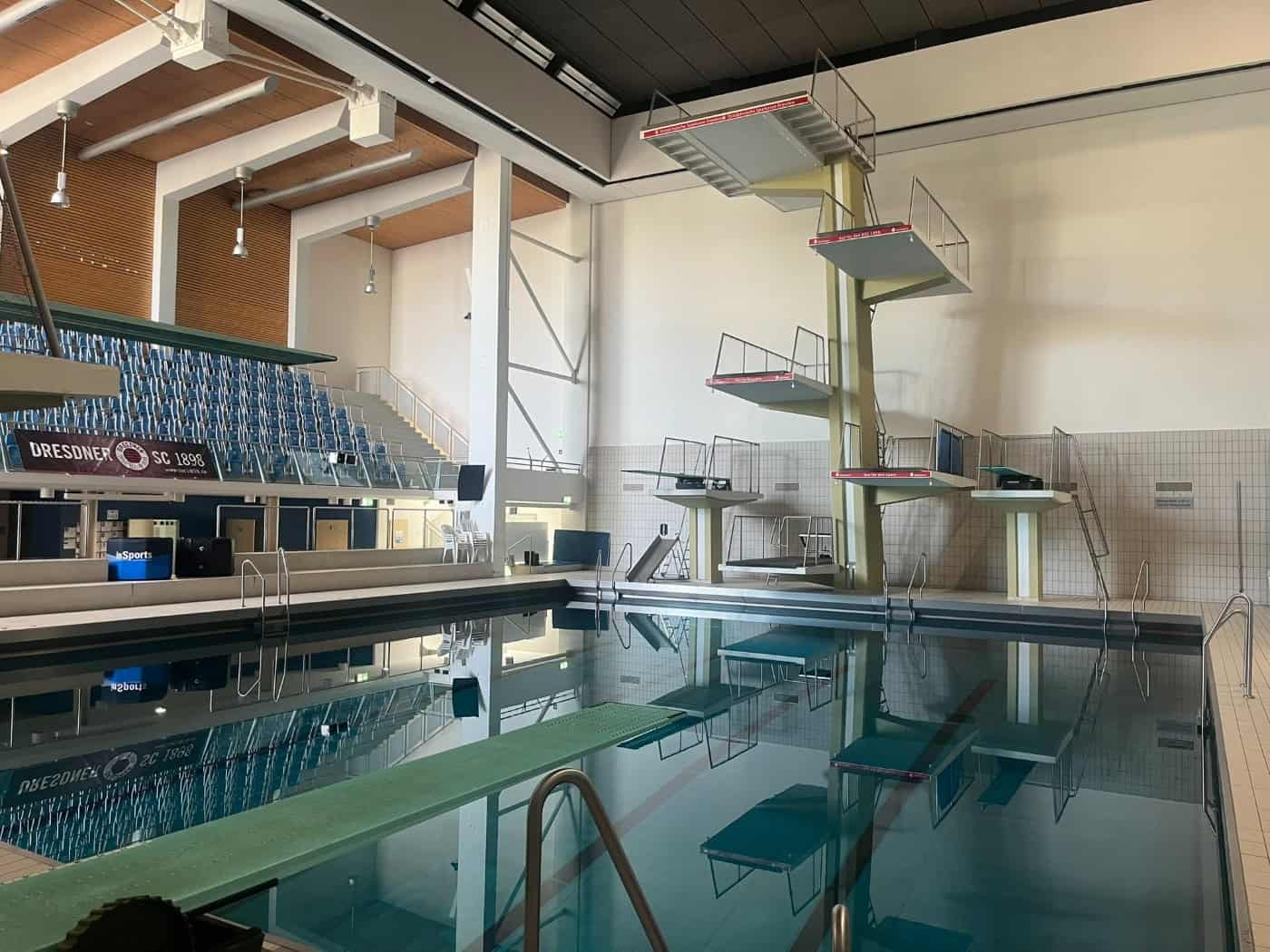 Großes Hallenschwimmbecken mit einem mehrstufigen Sprungturm, Sprungbreitern und einer blauen Zuschauertribüne im Hintergrund.