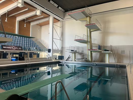 Großes Hallenschwimmbecken mit einem mehrstufigen Sprungturm, Sprungbreitern und einer blauen Zuschauertribüne im Hintergrund.