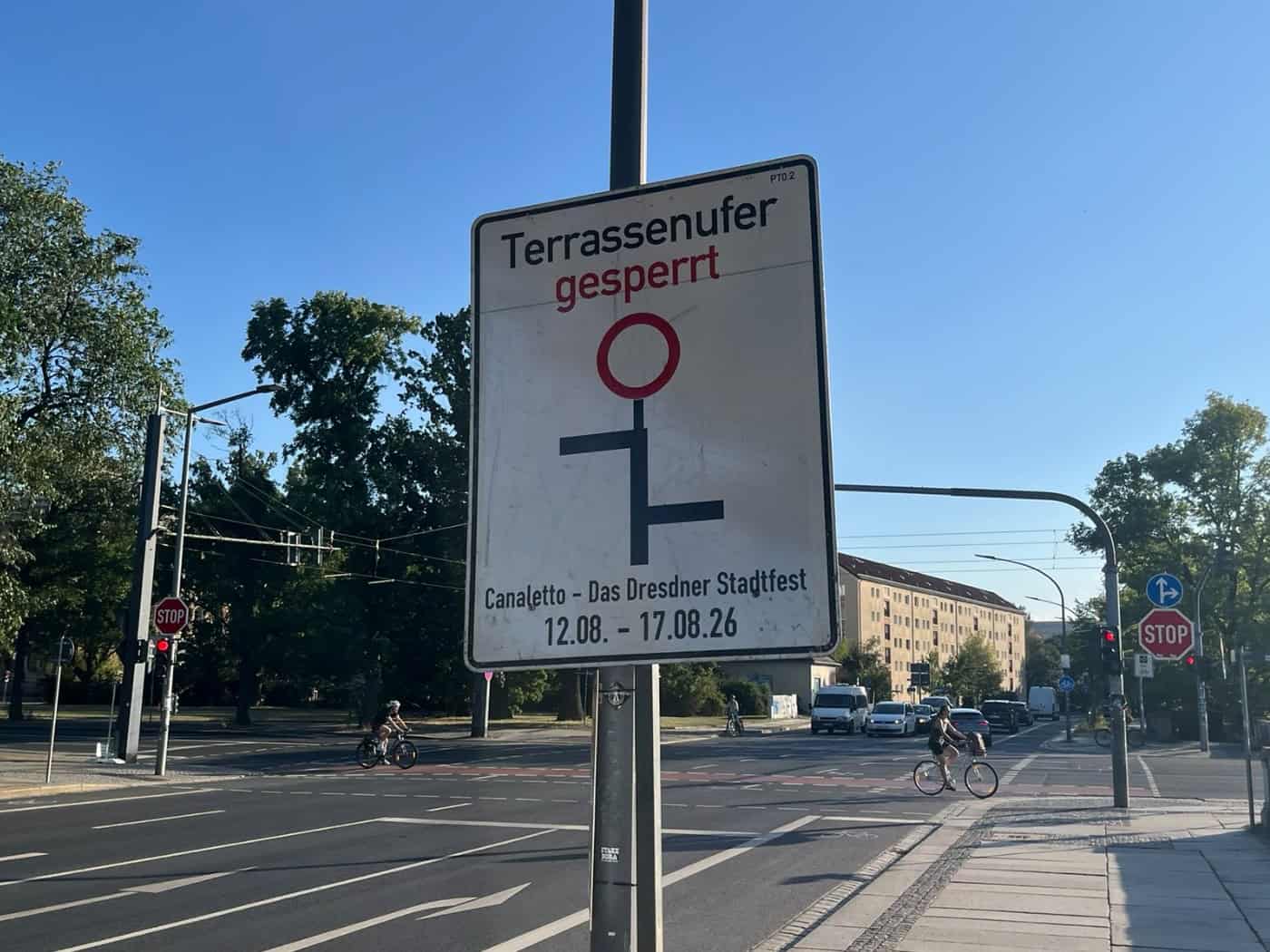 Weißes Hinweisschild an einer Straßenkreuzung in Dresden mit der Aufschrift „Terrassenufer gesperrt – Canaletto – Das Dresdner Stadtfest 12.08. – 17.08.26“. Im Hintergrund sind eine mehrspurige Straße, Bäume und Radfahrer bei sonnigem Wetter zu sehen.