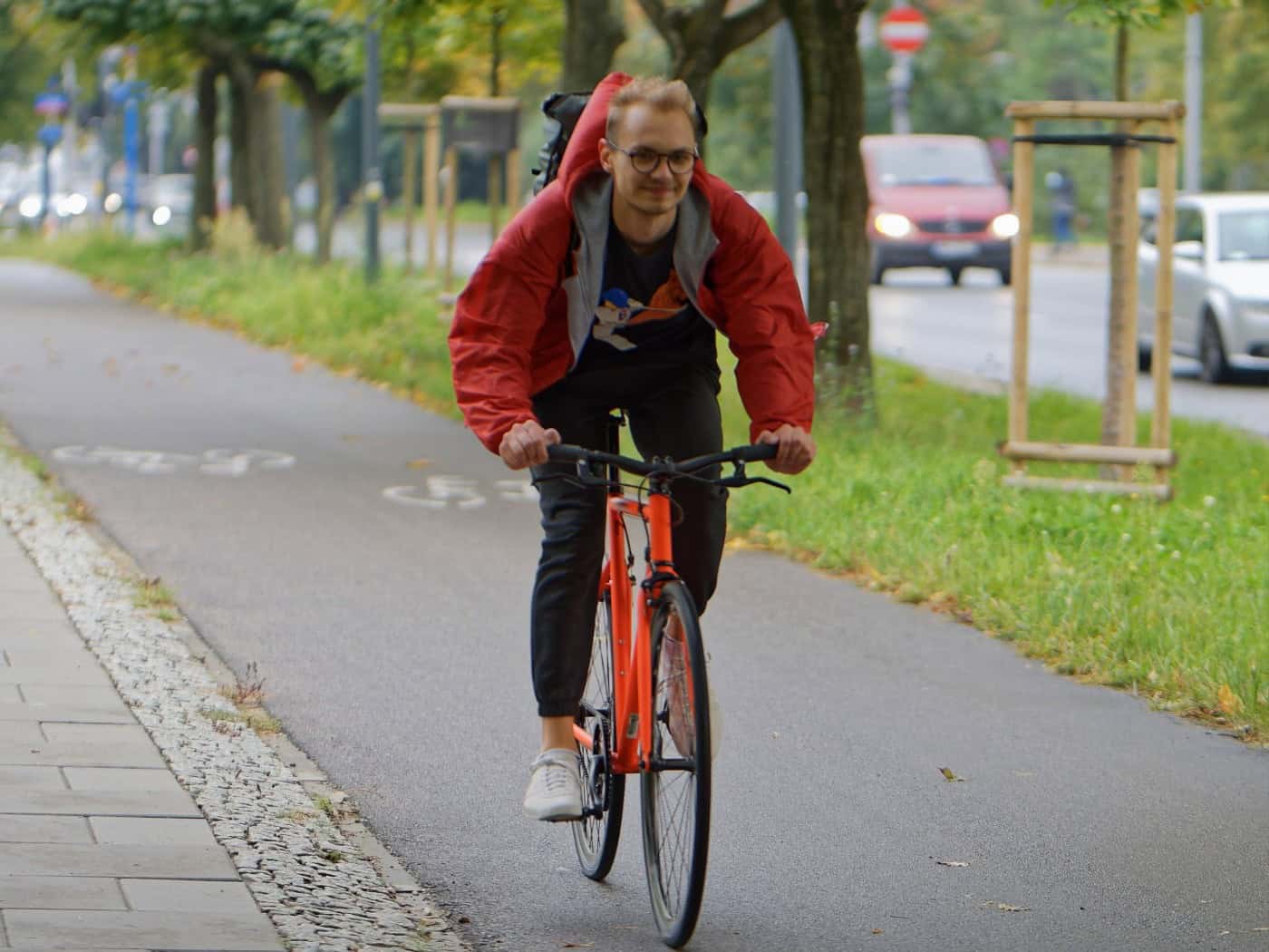 Ein junger Mann mit Brille und roter Jacke fährt auf einem orangefarbenen Fahrrad über einen asphaltierten Radweg neben einer Straße mit Bäumen.