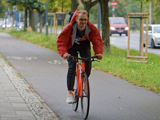 Ein junger Mann mit Brille und roter Jacke fährt auf einem orangefarbenen Fahrrad über einen asphaltierten Radweg neben einer Straße mit Bäumen.