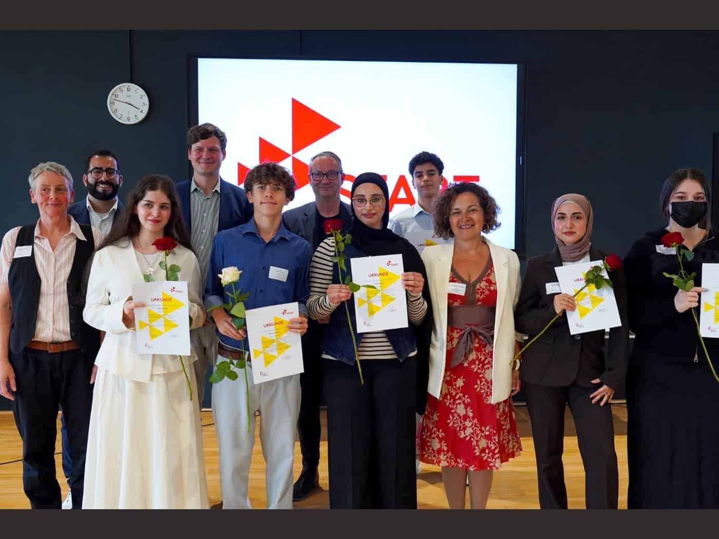 Eine Gruppe von Jugendlichen und Politikvertretern steht bei einer Festveranstaltung zusammen. Die Schülerinnen und Schüler halten stolz Urkunden und Rosen in den Händen. Im Hintergrund ist auf einem großen Bildschirm das rot-weiße START-Logo zu sehen.