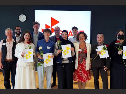 Eine Gruppe von Jugendlichen und Politikvertretern steht bei einer Festveranstaltung zusammen. Die Schülerinnen und Schüler halten stolz Urkunden und Rosen in den Händen. Im Hintergrund ist auf einem großen Bildschirm das rot-weiße START-Logo zu sehen.