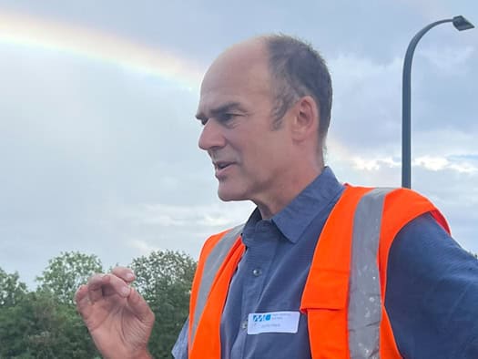 Prof. Steffen Marx mit Warnweste gestikuliert bei einem Pressetermin auf der Brücke Budapester Straße in Dresden. Im Hintergrund zeichnet sich vor bewölktem Himmel ein Regenbogen ab.